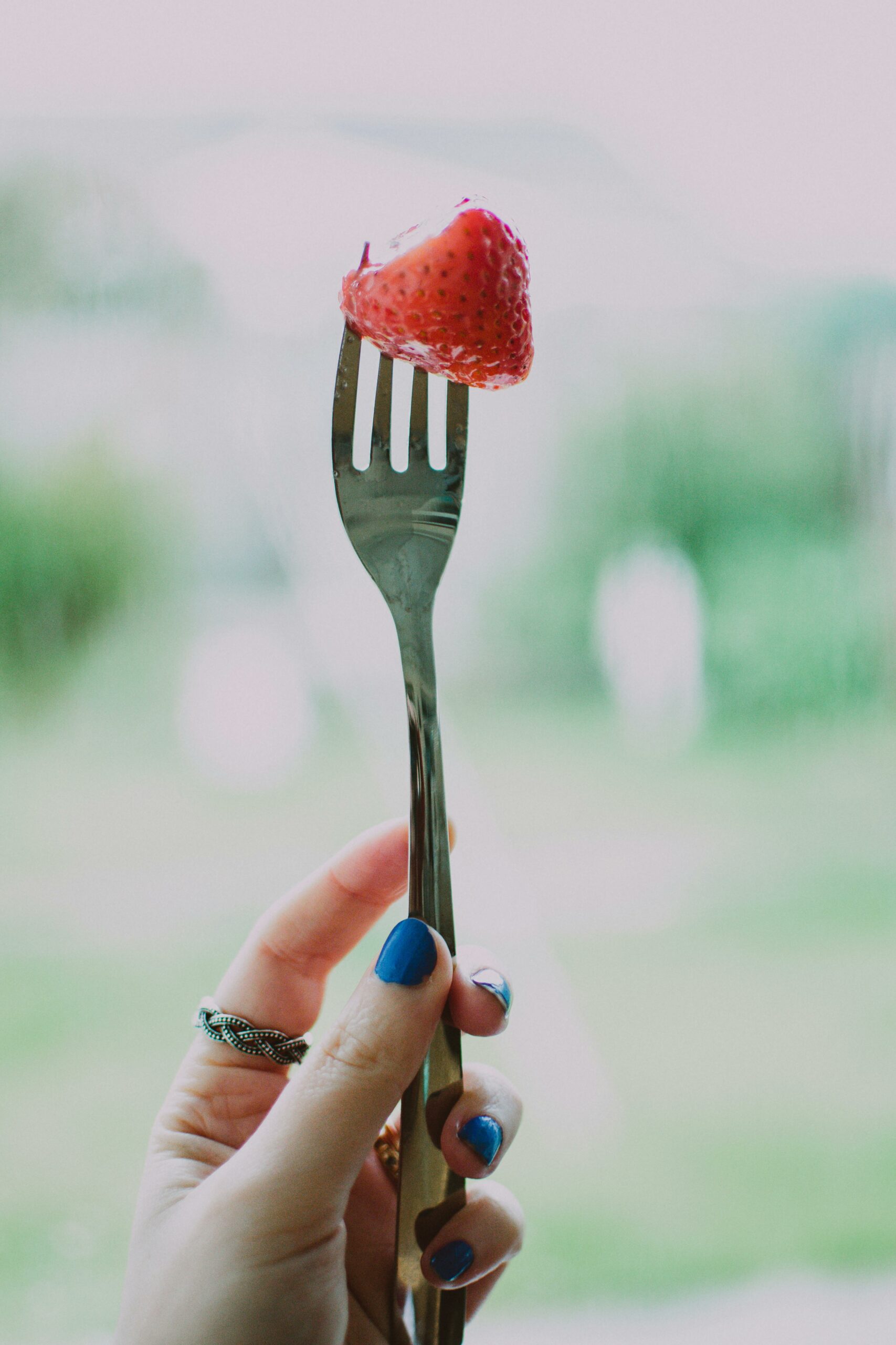 A hand with blue nail polish holds a fork with a fresh strawberry outdoors.